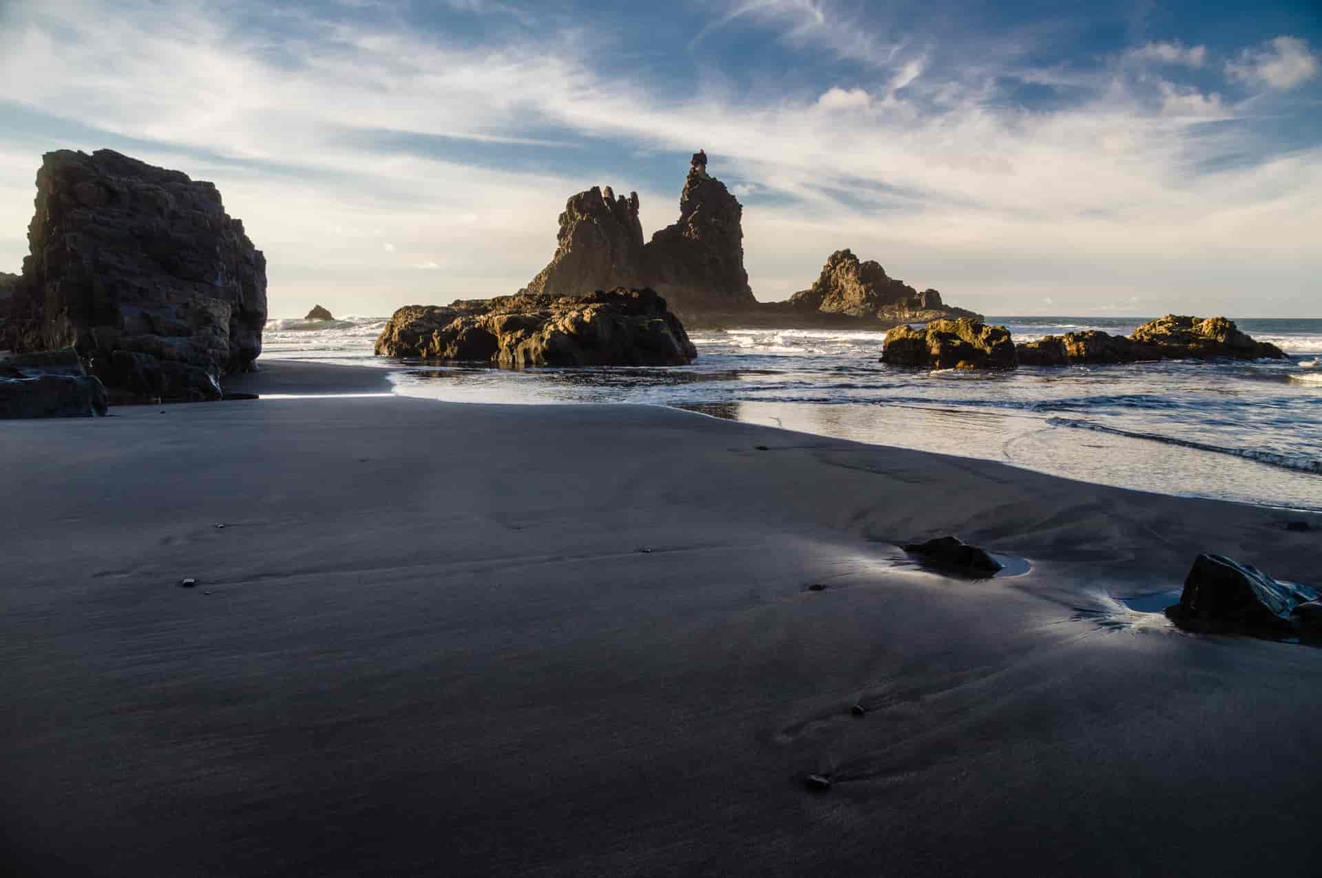 Rocky coast of Benijo beach with dark sand, sea stacks, and ocean waves under a cloudy sky.