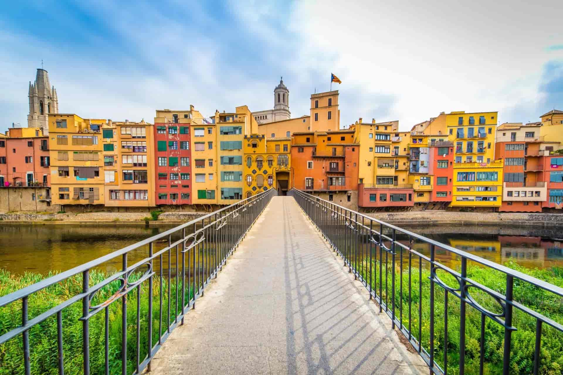 Footbridge leading to colorful riverside buildings and cathedral tower in Girona, Spain.