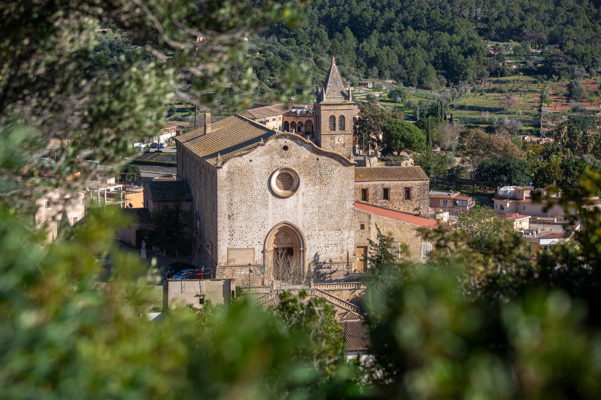 Stone church with bell tower seen through blurred green foliage in Andratx valley.