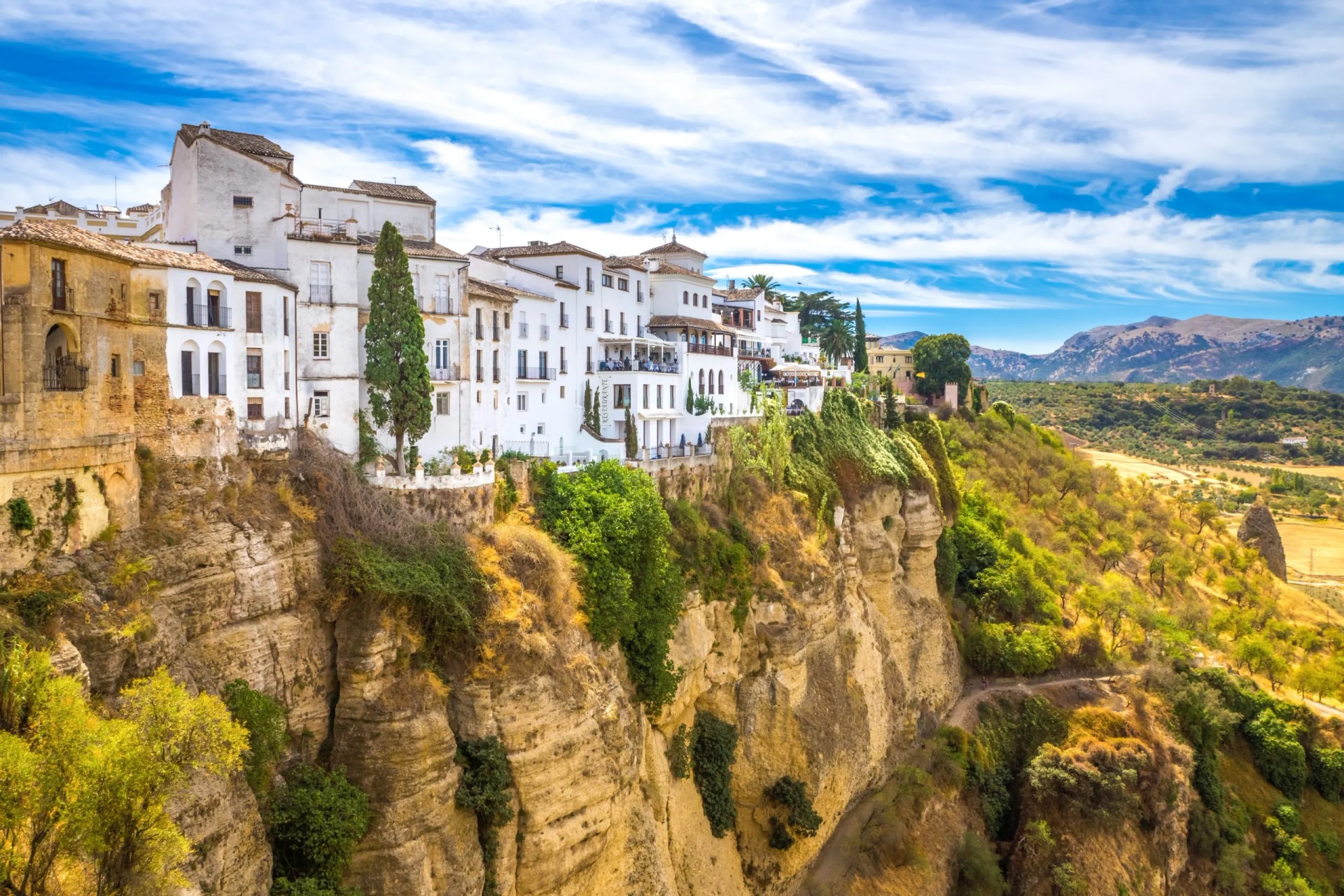 White buildings perched on a high cliff edge above lush green slopes under a blue sky in Ronda.
