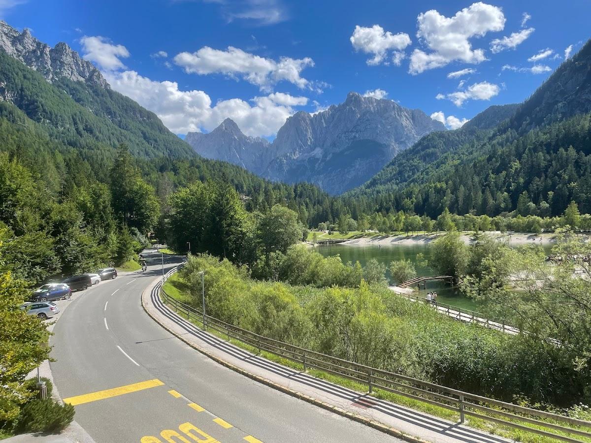 Winding road near a lake surrounded by lush green mountains under a blue sky with clouds.