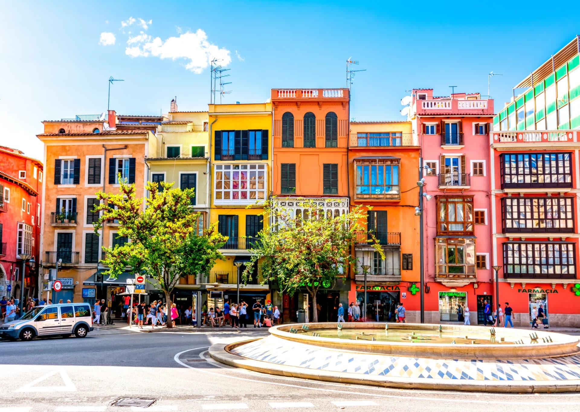Colorful buildings line a street with a fountain, people walking, and a van in Palma.