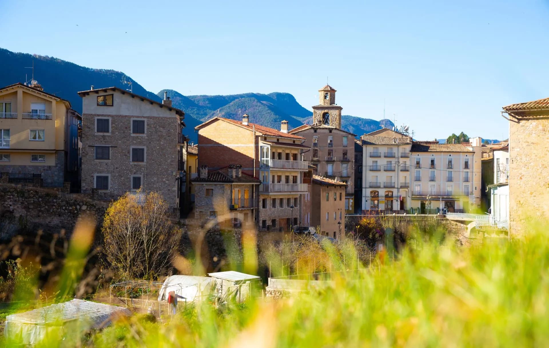 Townscape with stone buildings and bell tower in La Pobla de Lillet, Spain, seen over foreground grass.