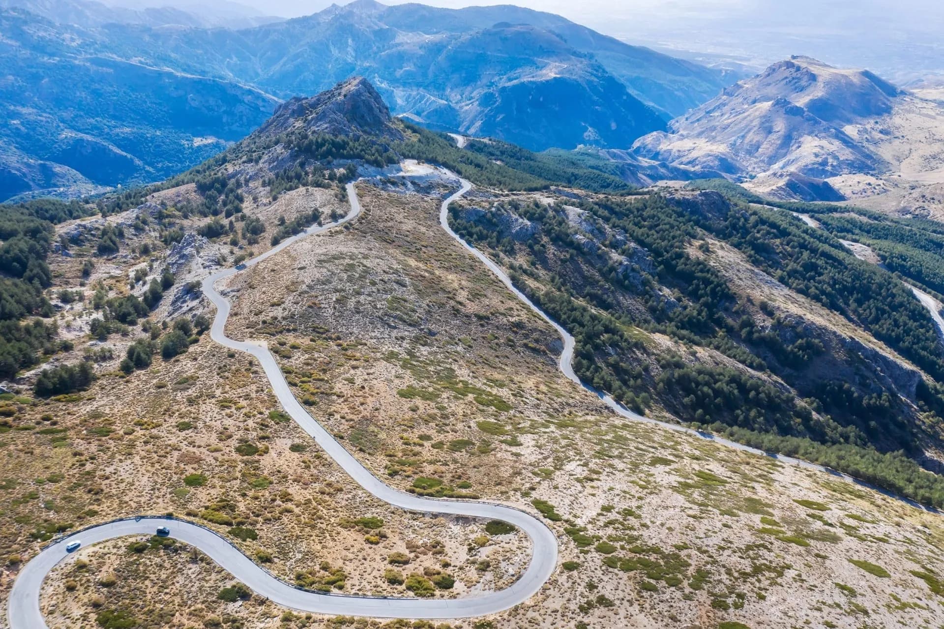 Winding mountain road near Sierra Nevada, Granada, Spain, with dry brush and distant blue peaks.
