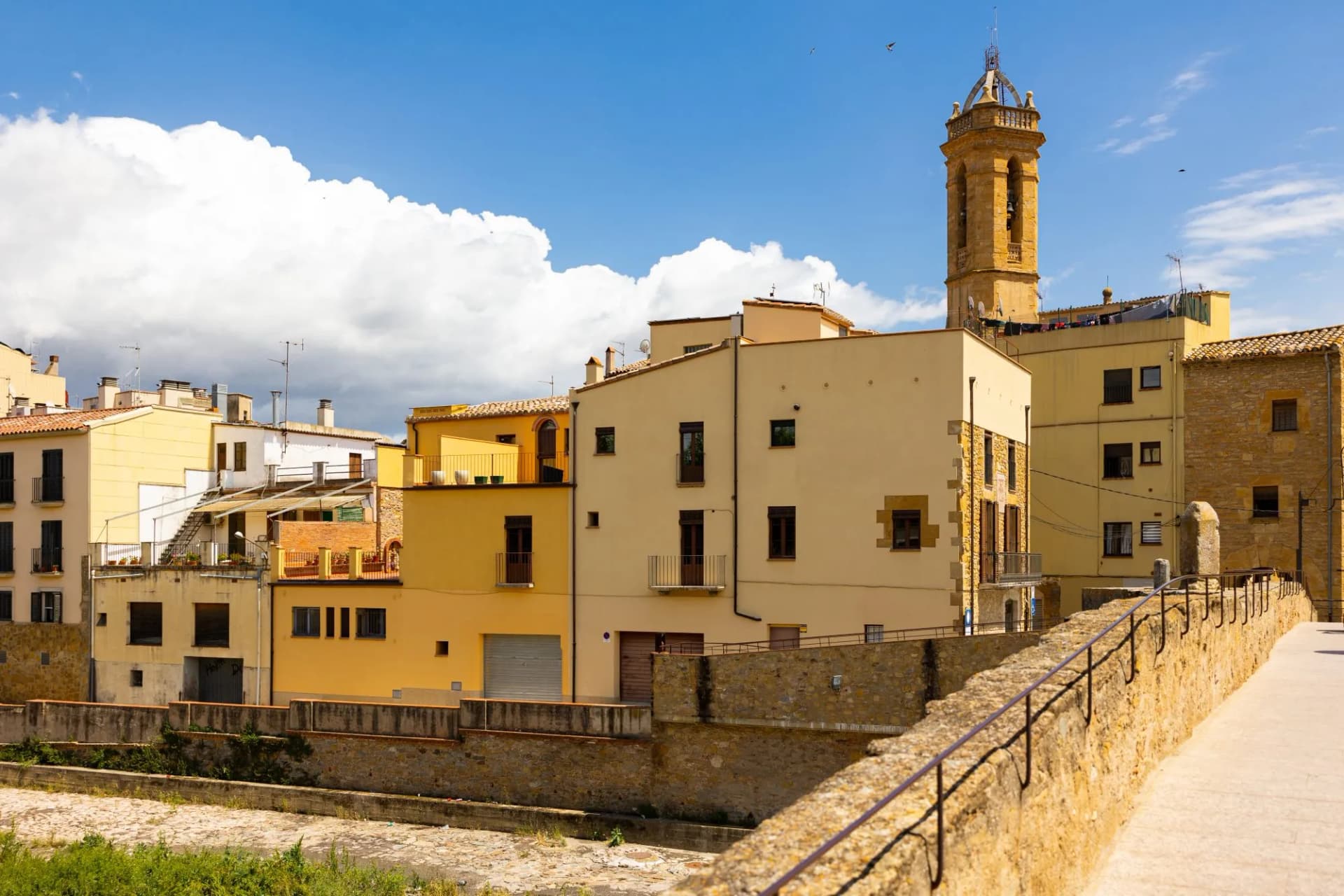 Village buildings with yellow facades and a stone bell tower under a blue sky in La Bisbal d'Empordà.