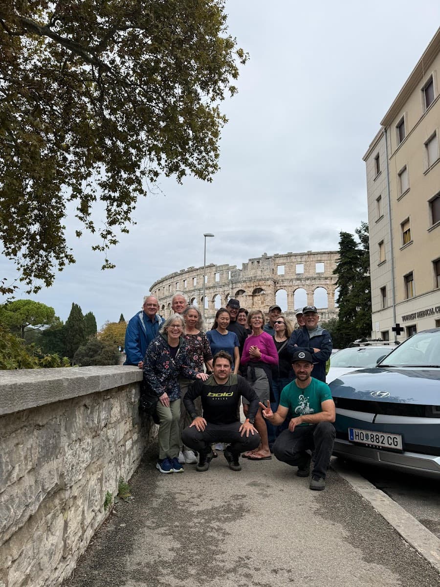 Group posing in front of Pula Arena with a stone wall and parked car.