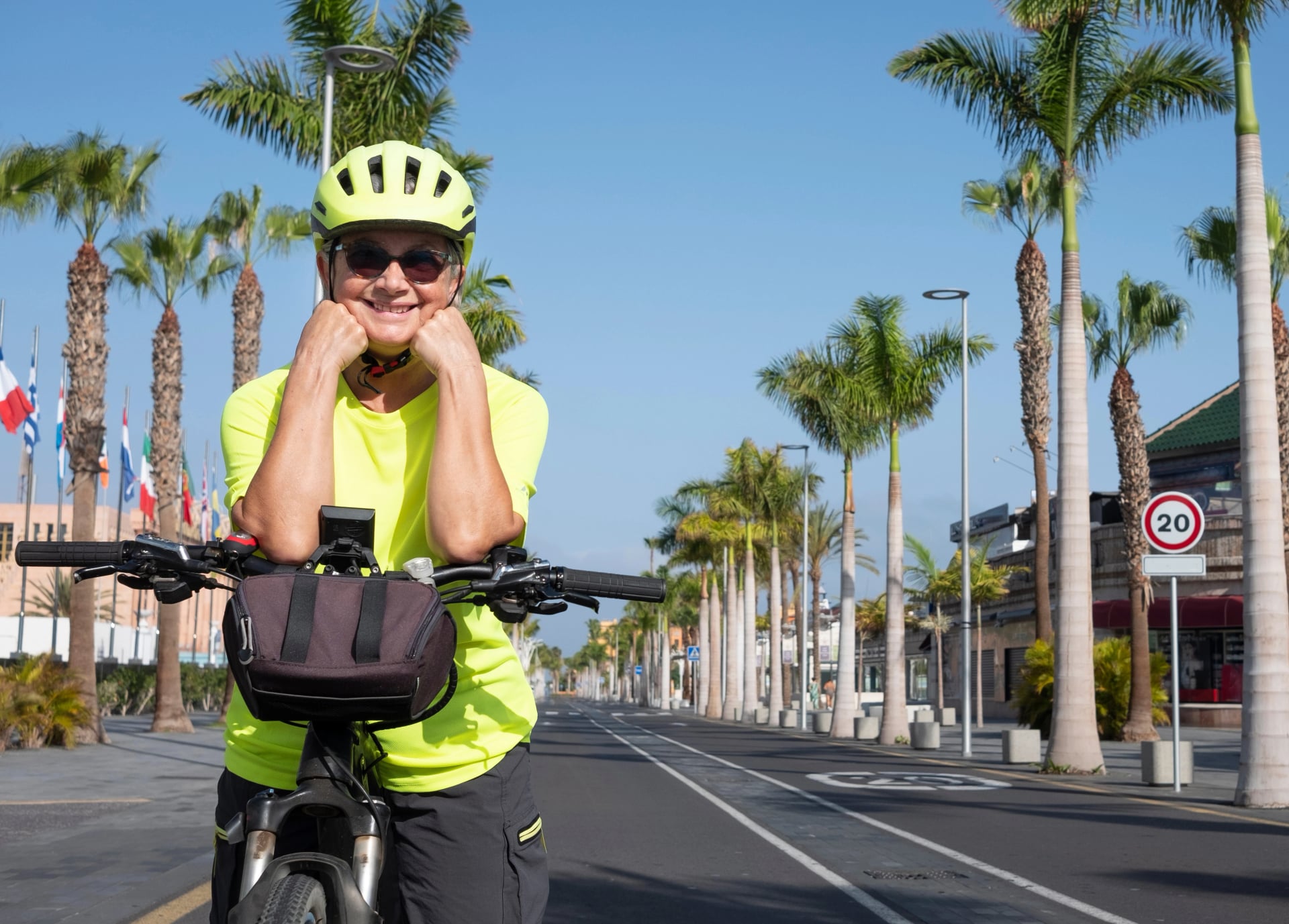 Aktiv smilende senior kvinde med briller og gul hjelm, der cykler på den øde vej Tenerife, De Kanariske Øer. Palmetræer og blå himmel i baggrunden