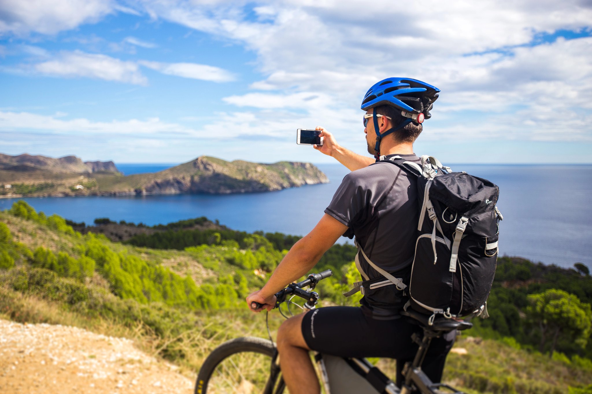 ung kille på mountainbikeleder i Spanien och tar foto med vit telefon i bakgrunden av Medelhavet vid den klippiga kusten Costa Brava. I blå hjälm på huvudet