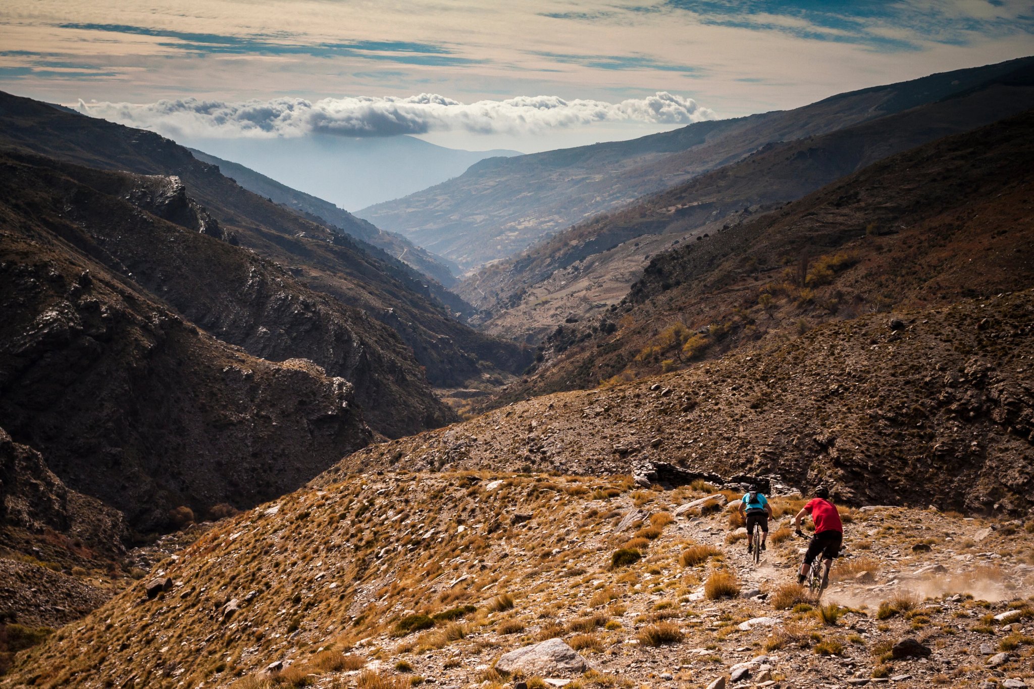 SIERRA NEVADA, SPANIA. To terrengsyklister som sykler ned en grov singletrack sti i en øde, avsidesliggende dal med dramatisk himmel og tåke.